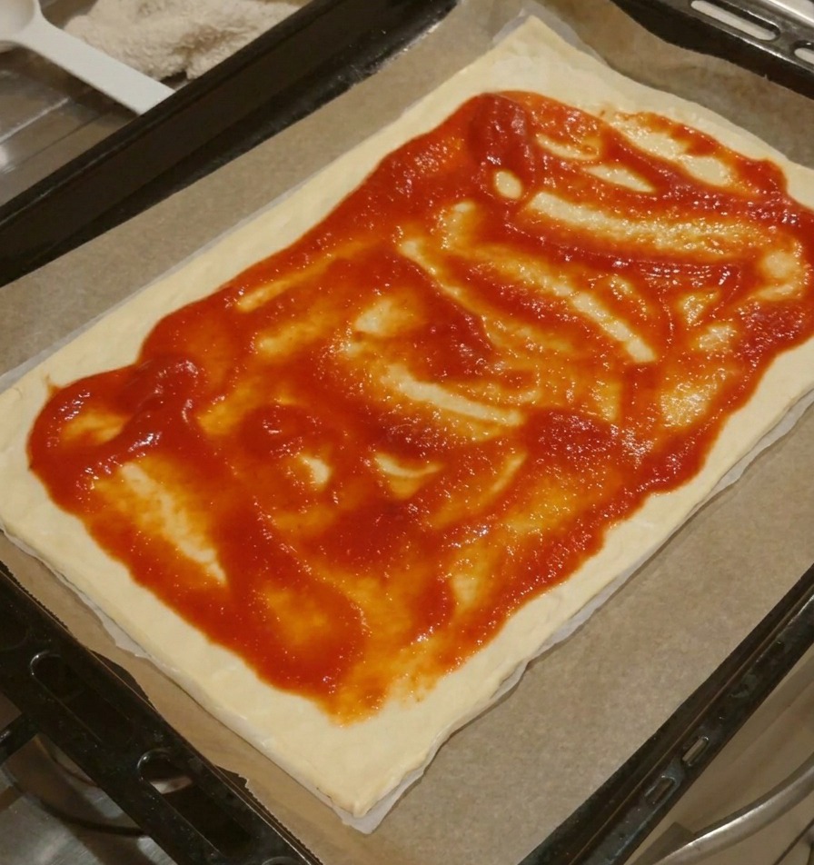 A rectangular pizza base with red tomato sauce spread evenly across the surface, leaving a clear dough border.