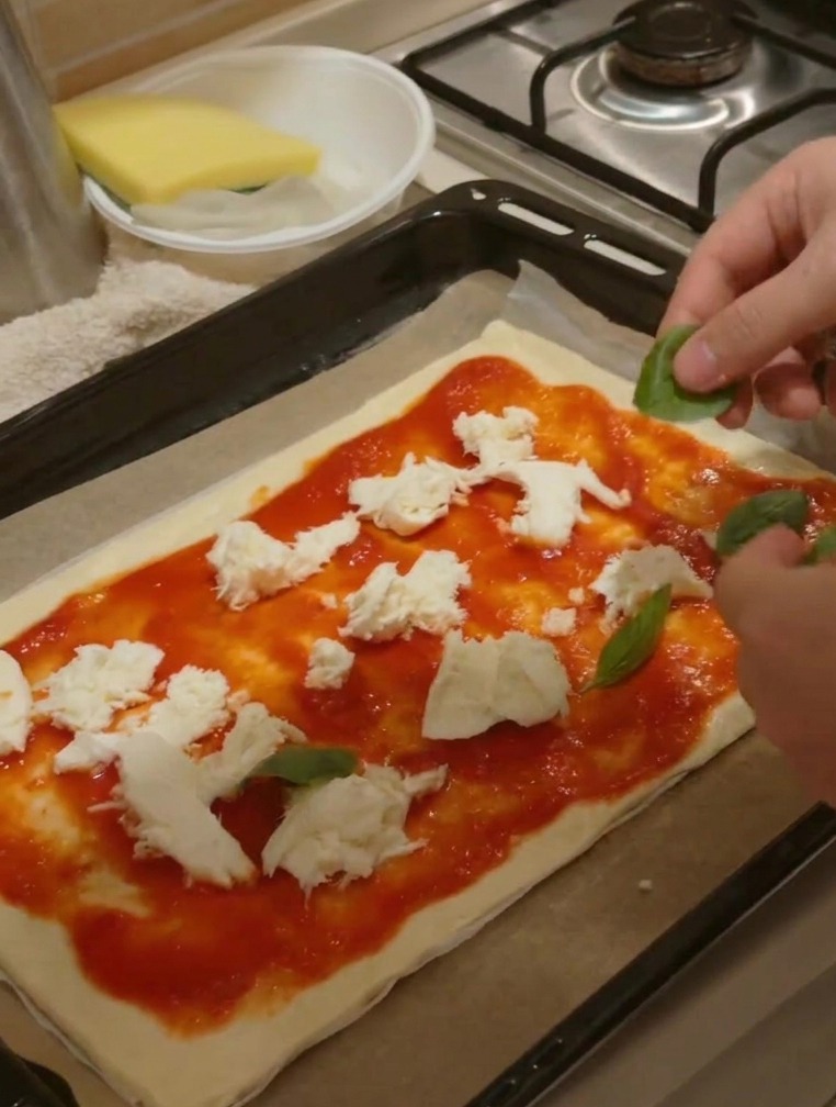 Hands placing fresh green basil leaves onto a pizza already topped with sauce and cheese chunks.