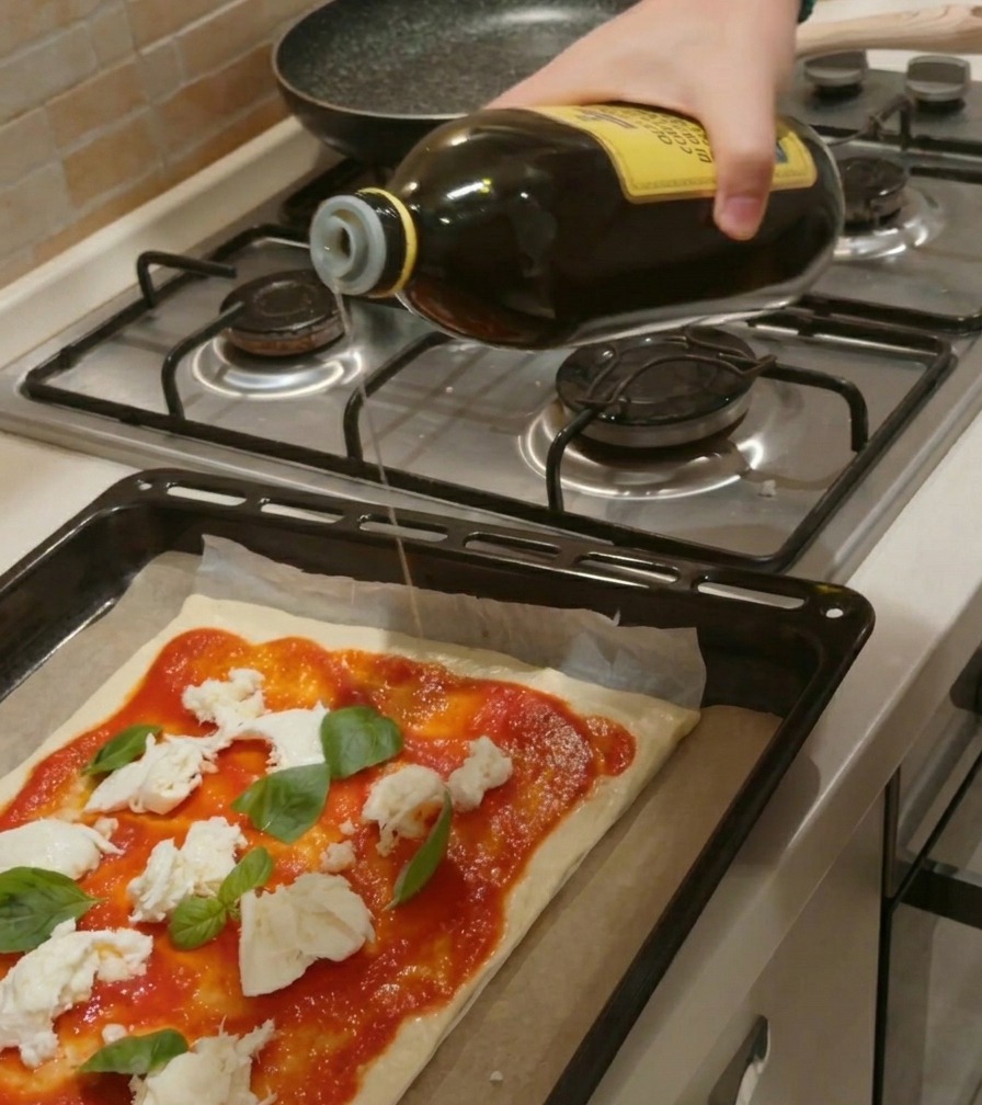 A hand pouring a stream of olive oil from a large glass bottle onto a raw pizza crust topped with red tomato sauce, chunks of white mozzarella, and green basil leaves.