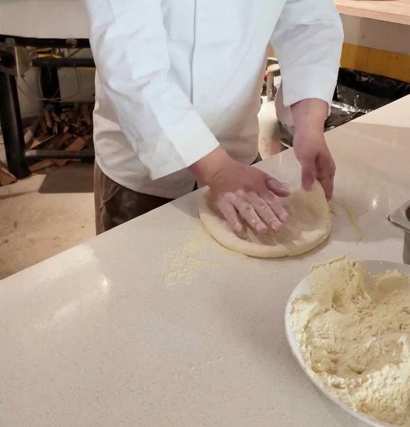 A chefs hands stretching a round piece of raw pizza dough on a floured countertop in front of a wood-fired oven.