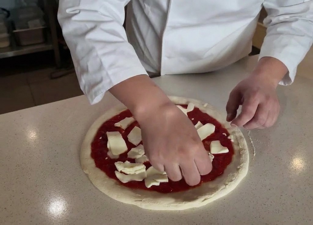 A chefs hands placing torn pieces of fresh white mozzarella cheese onto a pizza base spread with red tomato sauce.