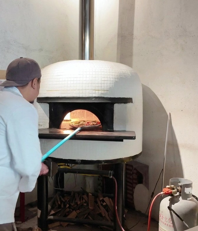 A pizza being launched from a long handled peel into the mouth of a white tiled glowing wood fired pizza oven.