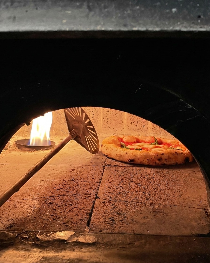 A Margherita pizza cooking inside a wood fired stone oven being rotated by a small metal peel near the fire.