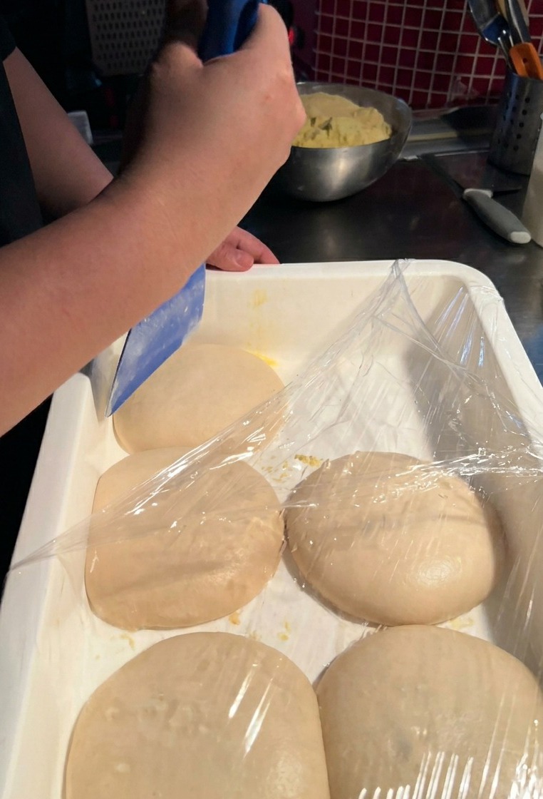 A person using a blue plastic scraper to carefully lift a smooth, round portion of pizza dough from a white plastic container.