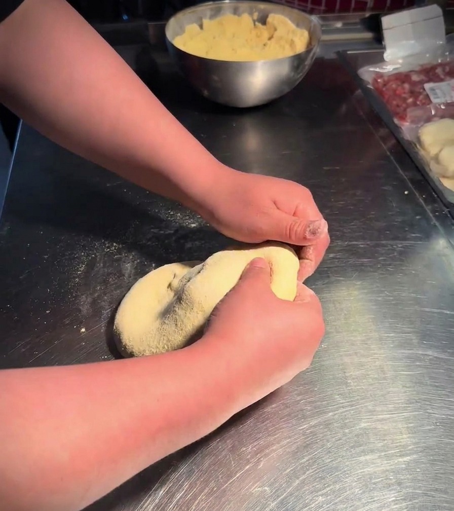 Two hands pressing and flipping a soft dough ball in a stainless steel bowl filled with yellow cornmeal.