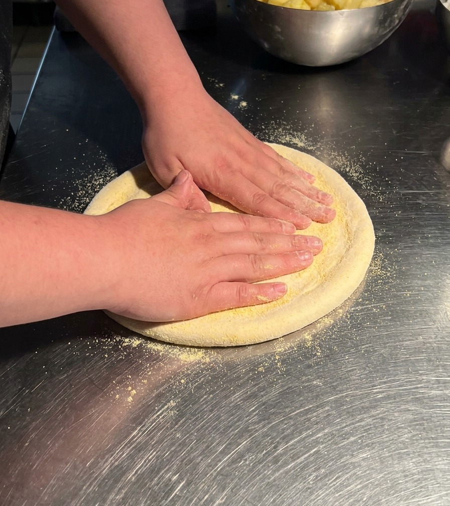 A persons hands pressing down on a circular piece of pizza dough on a stainless steel table, stretching it into a larger disk.