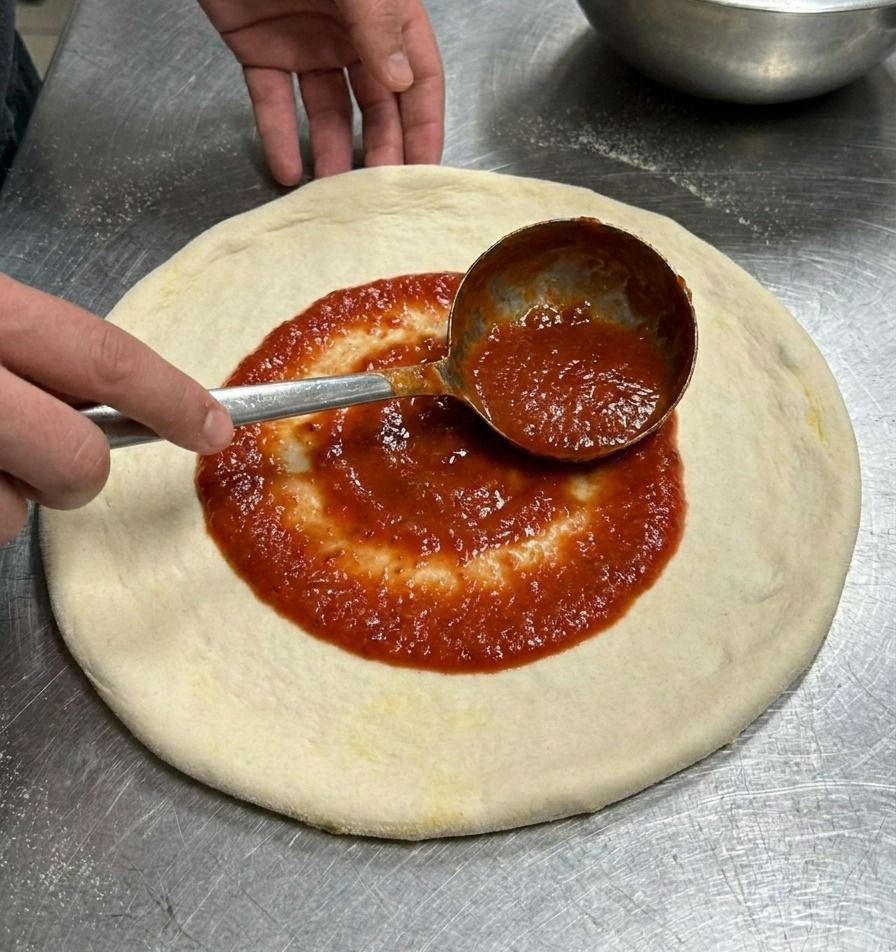 A metal ladle spreading vibrant red tomato sauce in a circular motion over a flat, round pizza dough.