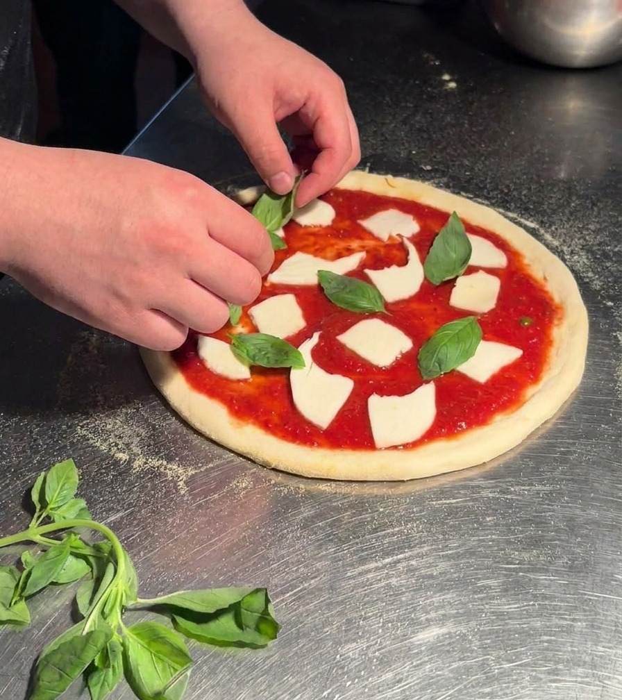 Hands placing fresh, bright green basil leaves onto a raw Margherita pizza prepared with sauce and mozzarella.