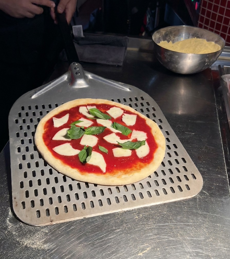 A raw Margherita pizza topped with tomato sauce, fresh mozzarella, and basil leaves resting on a large, perforated metal pizza peel.