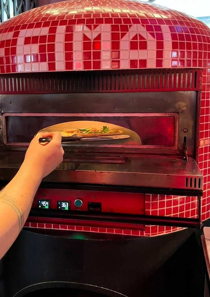 A person sliding a raw pizza from a metal peel into the glowing mouth of a traditional red-tiled pizza kiln.