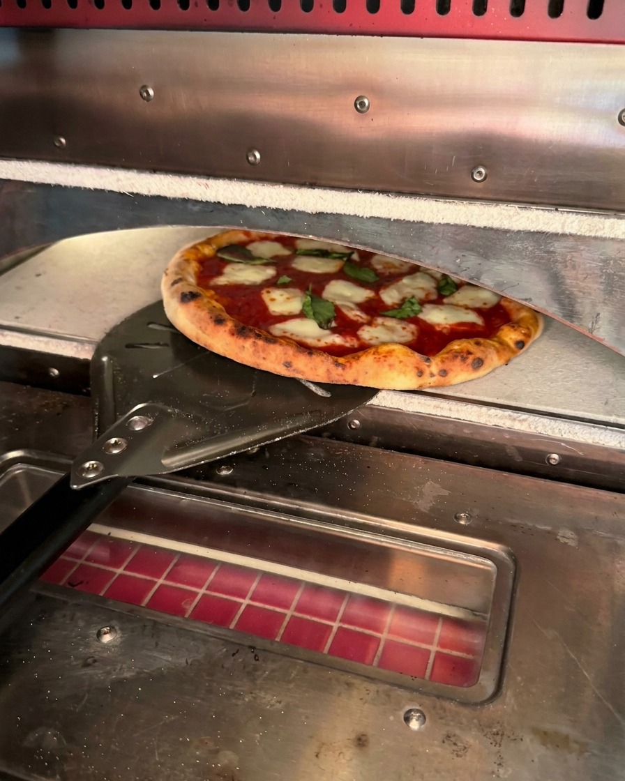 A freshly baked pizza with charred crust edges being retrieved from a pizza oven using a metal peel.