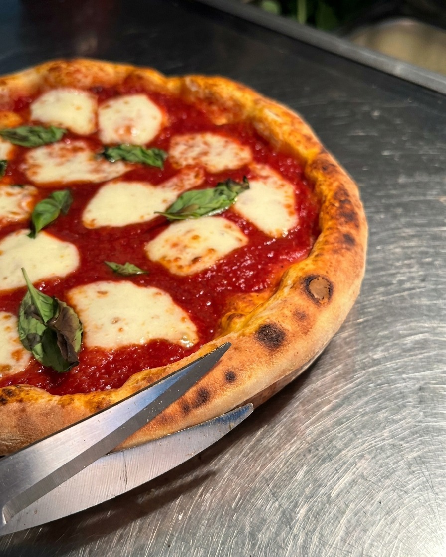 A close-up shot of sharp metal kitchen scissors cutting through the puffed crust of a hot Margherita pizza.