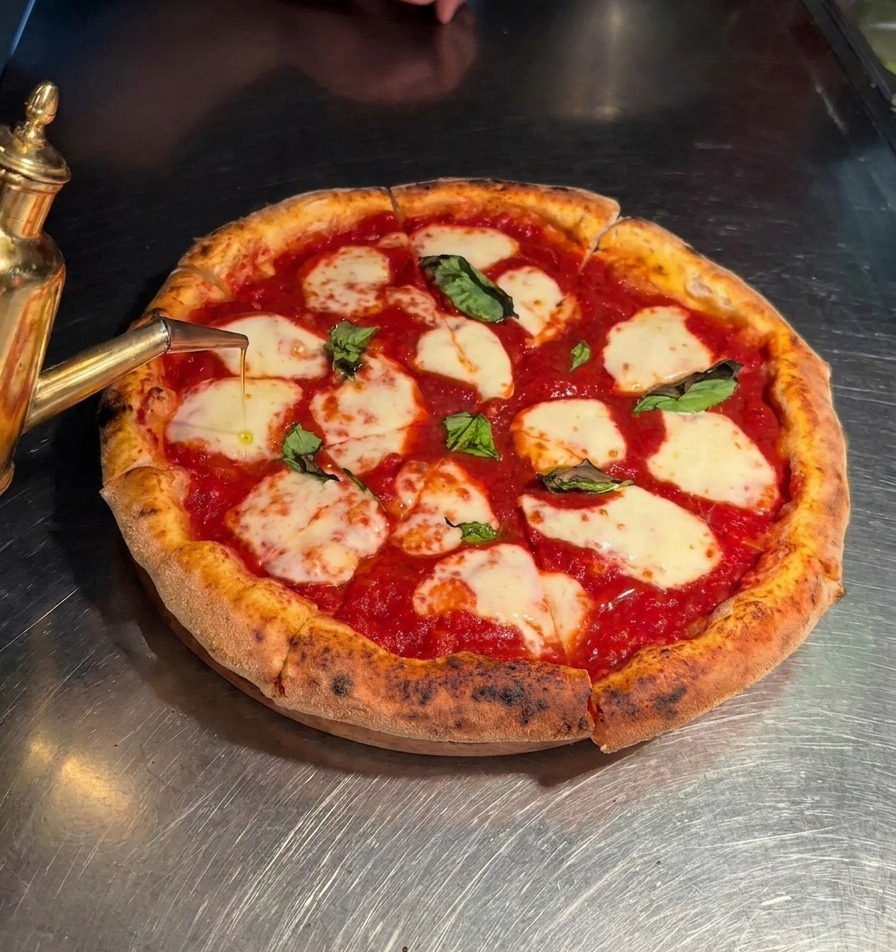 Extra virgin olive oil being poured from a golden spouted dispenser onto a sliced Margherita pizza.