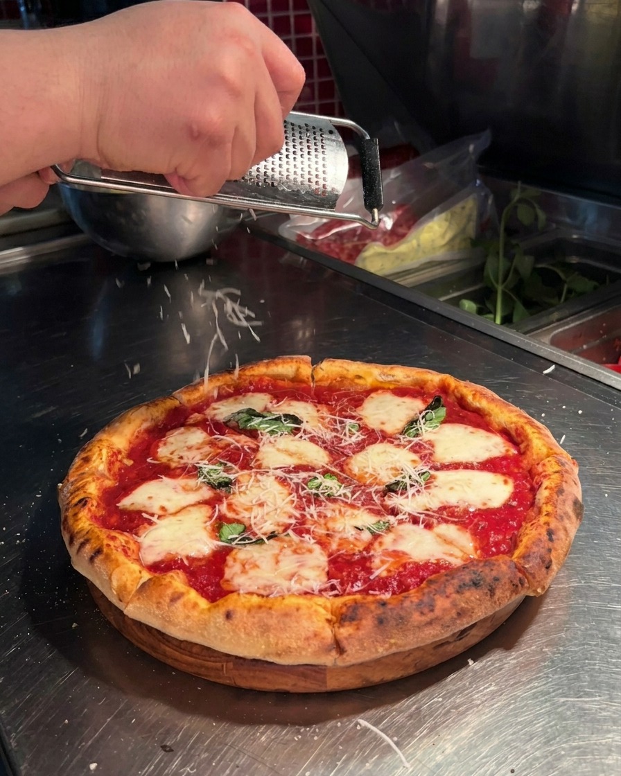 A close-up shot of a hand using a microplane to grate fresh parmesan cheese over a Neapolitan-style pizza topped with tomato sauce, mozzarella, and basil.