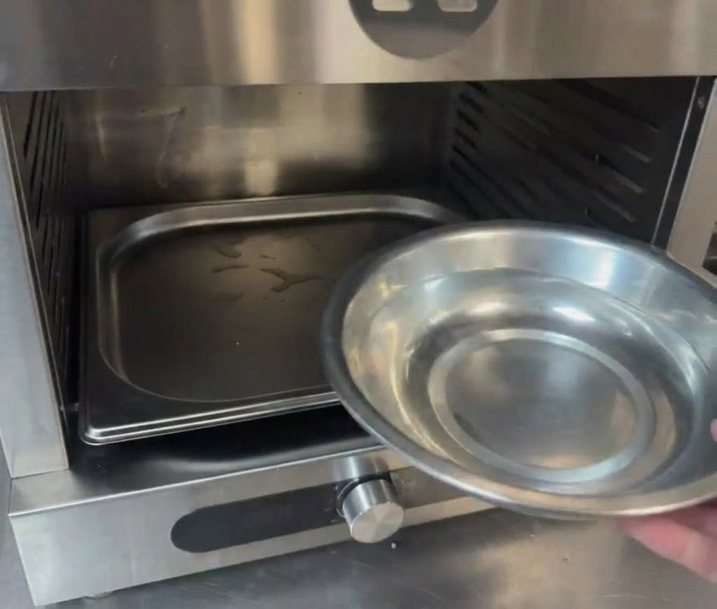 A hand pouring water from a small metal bowl into a tray located at the bottom of a professional oven chamber.