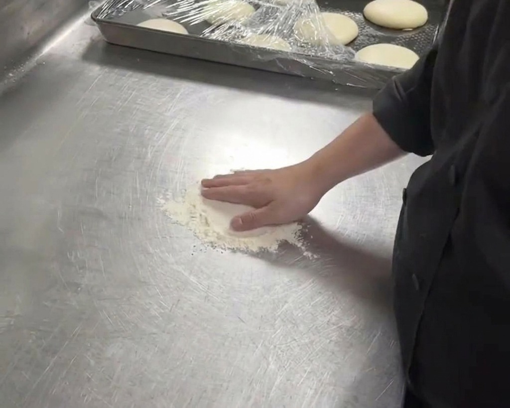 A person in a black chefs coat pressing their hand into a white dough ball on a floured metal counter.