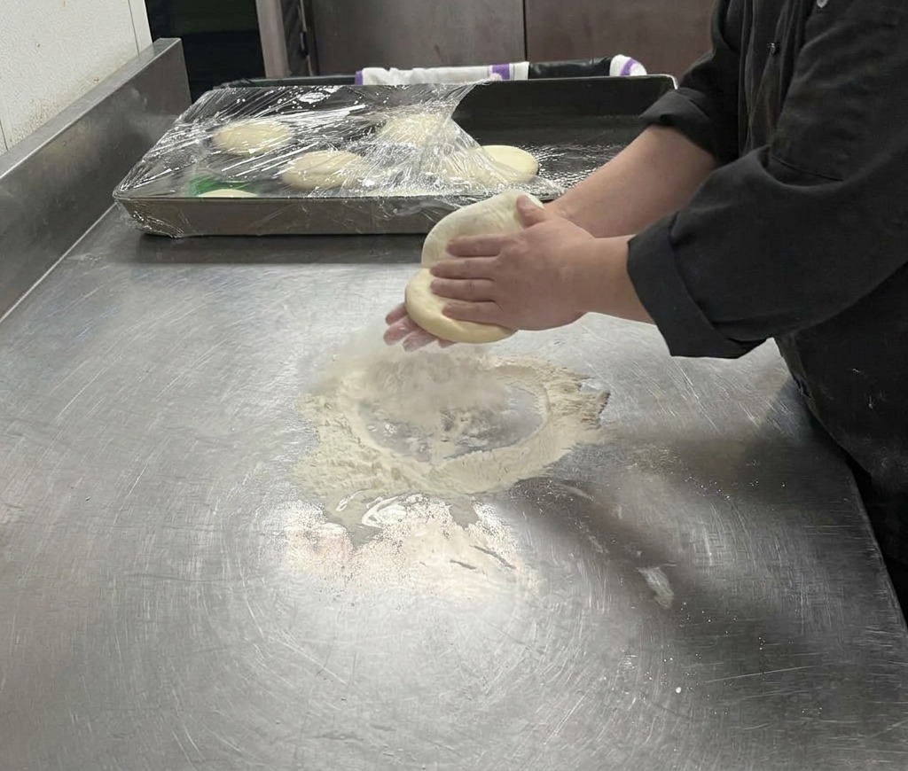Hands stretching a pale pizza dough over a floured surface, pulling it into a larger circle.