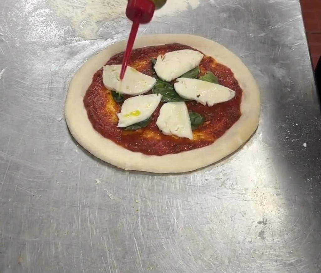 A red squeeze bottle drizzling extra virgin olive oil over a pizza topped with tomato sauce, basil, and mozzarella cheese.