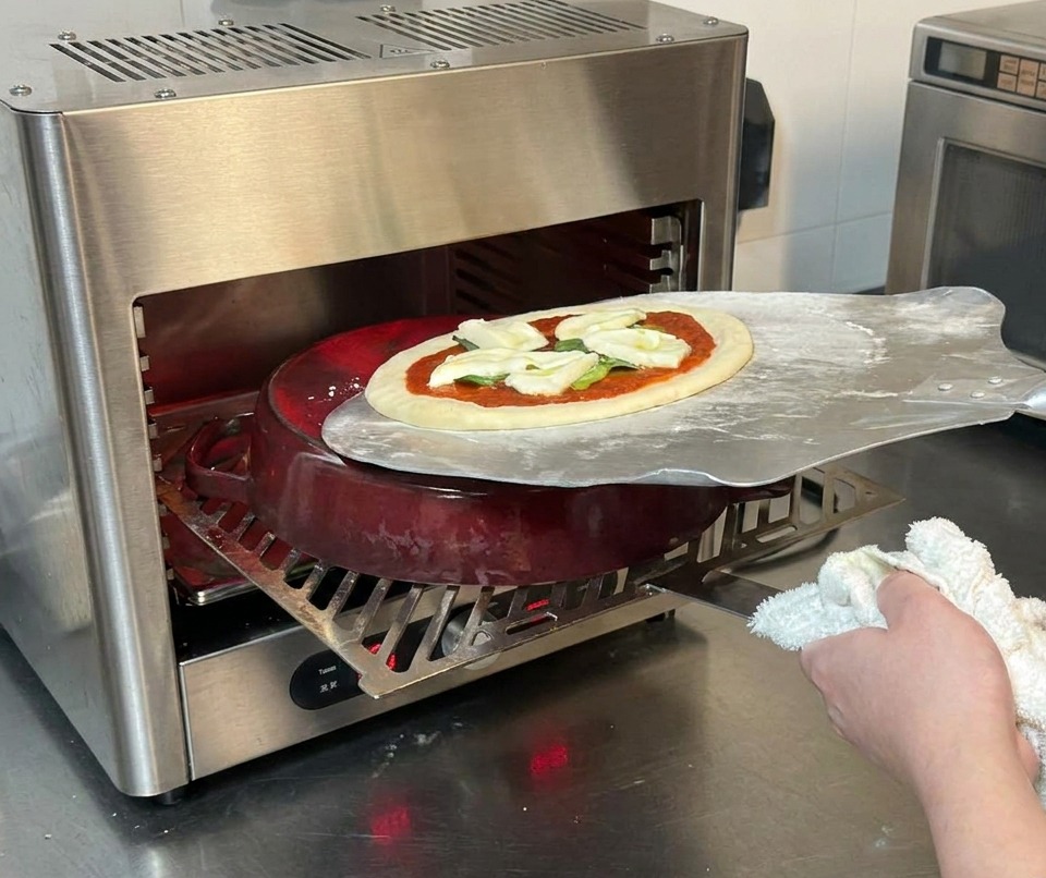 A metal pizza peel sliding an unbaked pizza onto a red cast iron pot inside a high temperature stainless steel oven.