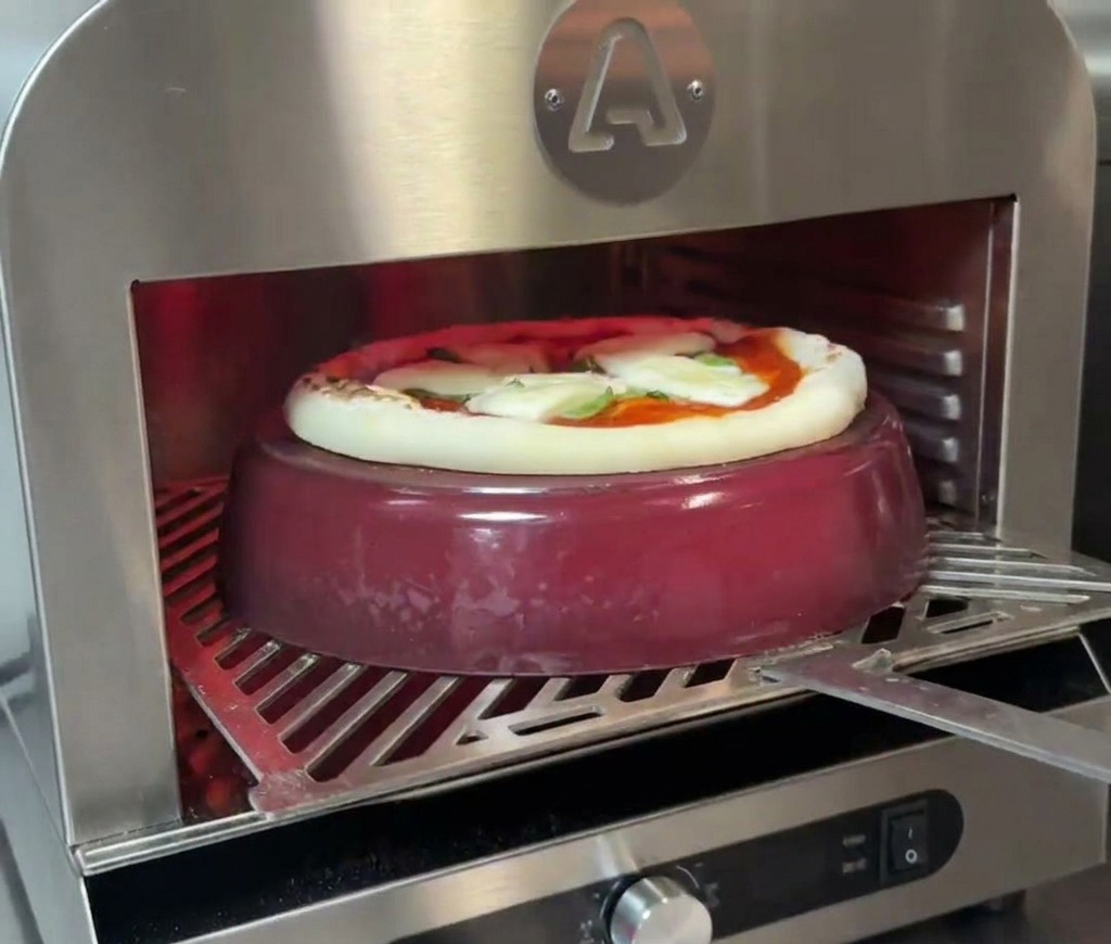 A pizza baking on top of a red cast iron pot inside an oven, with the crust visibly beginning to rise and brown.