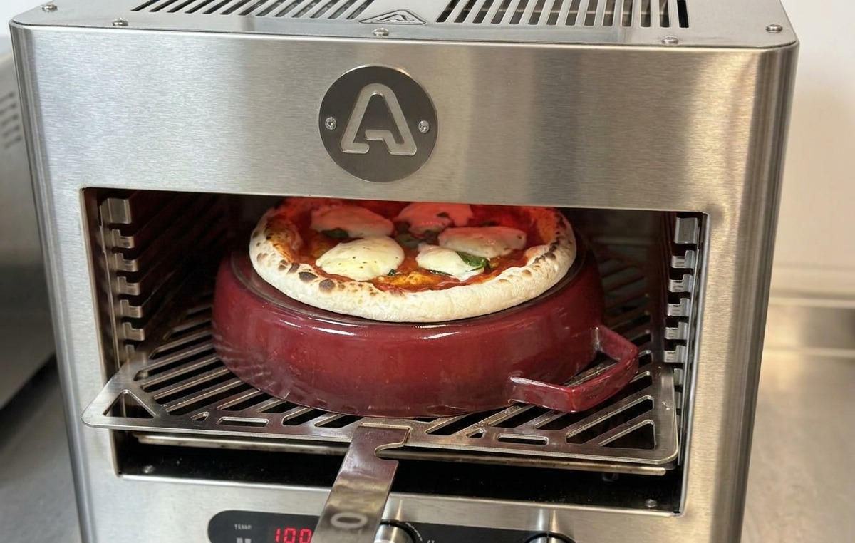 A metal pizza peel inserted into the oven to rotate a pizza that is nearly finished baking on a red cast iron pot.