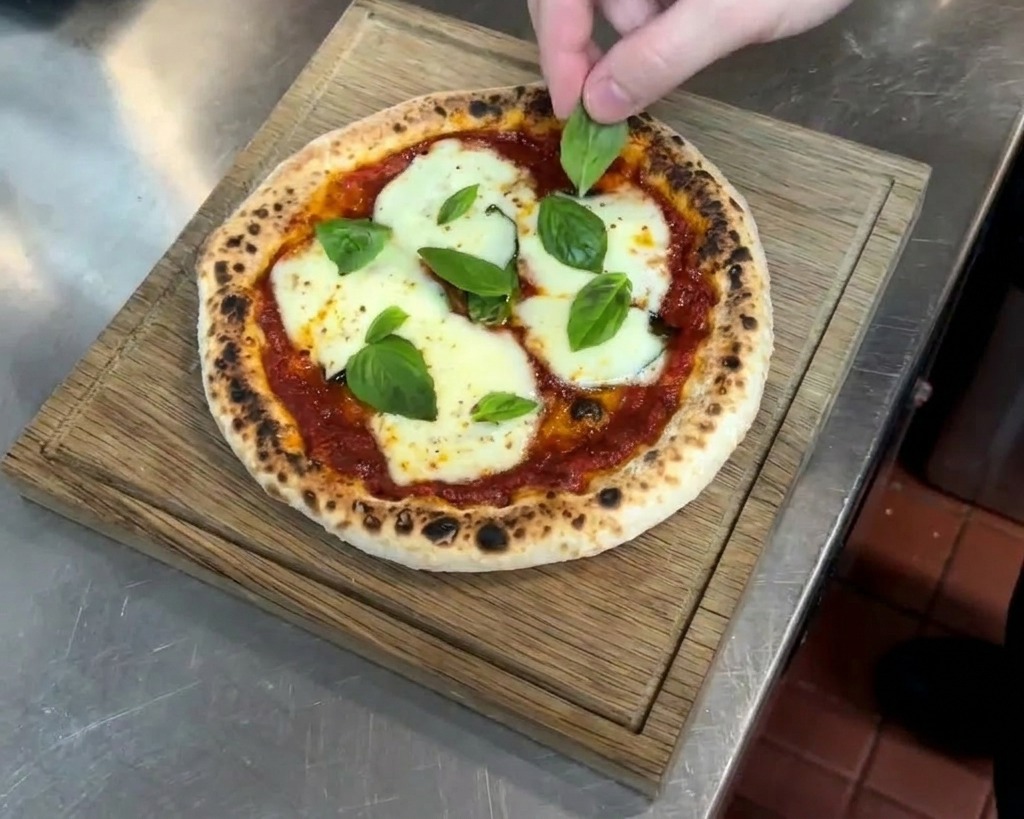 A persons hand carefully placing fresh green basil leaves onto a hot, freshly baked pizza with melted white cheese and a spotted, charred crust.