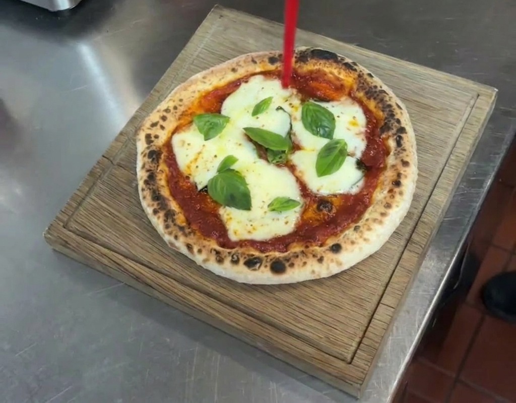 A stream of extra virgin olive oil being drizzled from a red tipped squeeze bottle onto a finished Margherita pizza resting on a wooden board.