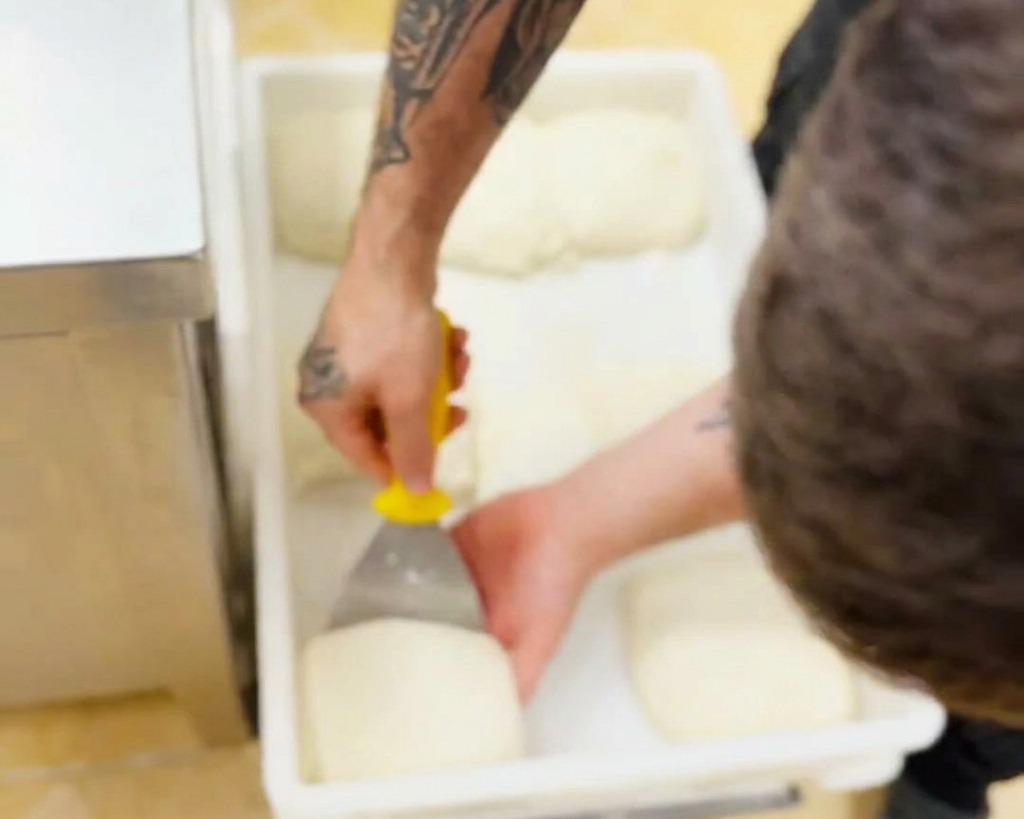 A bakers hand using a yellow-handled scraper to carefully lift a soft, white dough ball out of a rectangular plastic container.