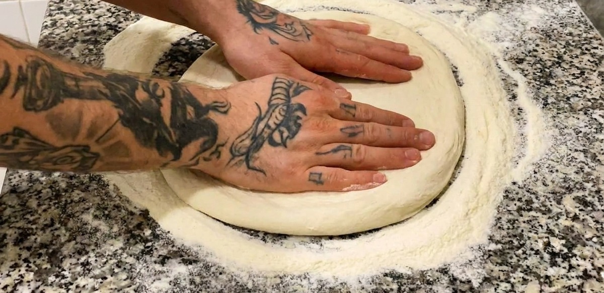 Two hands with tattoos pressing down on a round, floured pizza dough base on a grey marble countertop.