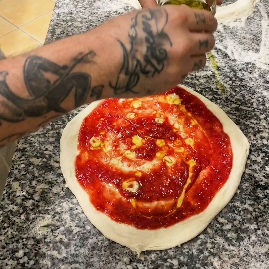 A hand using a metal spoon to spread bright red tomato sauce in a spiral pattern over raw pizza dough