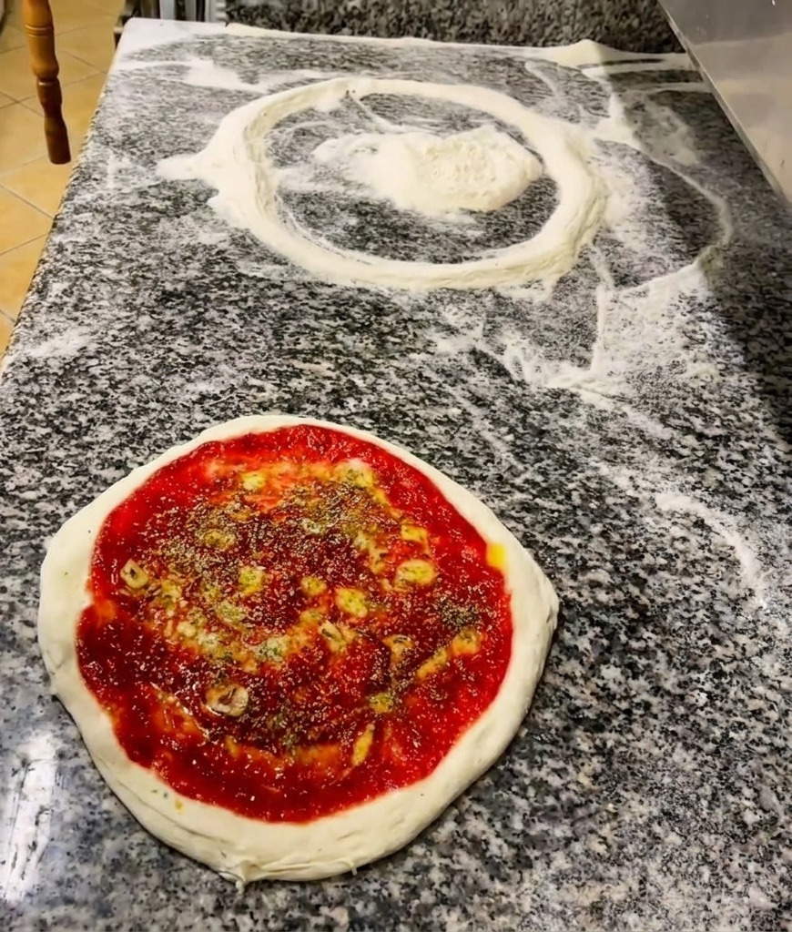A hand sprinkling green dried herbs over a pizza base already coated with tomato sauce and a drizzle of oil