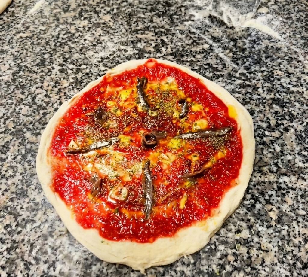 An overhead shot of an unbaked pizza base with tomato sauce, sprinkled herbs, and several silver anchovy fillets arranged like spokes on a wheel.