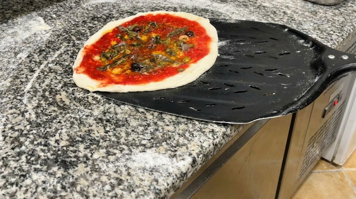 A seafood pizza ready for baking, resting on a perforated black metal pizza peel on a granite countertop.