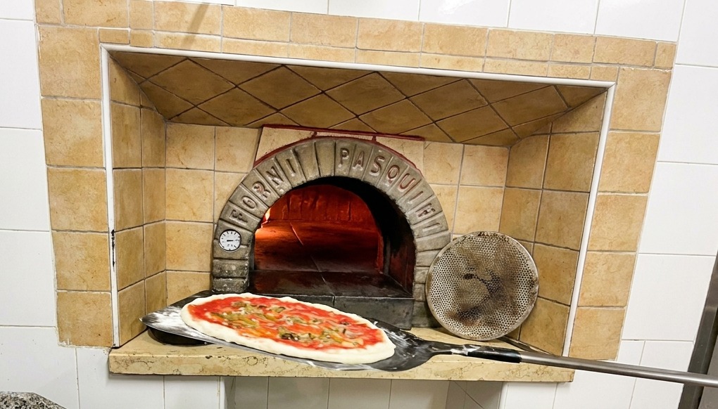 A pizza on a long-handled peel being placed into a traditional brick wood-fired oven with bright orange flames.