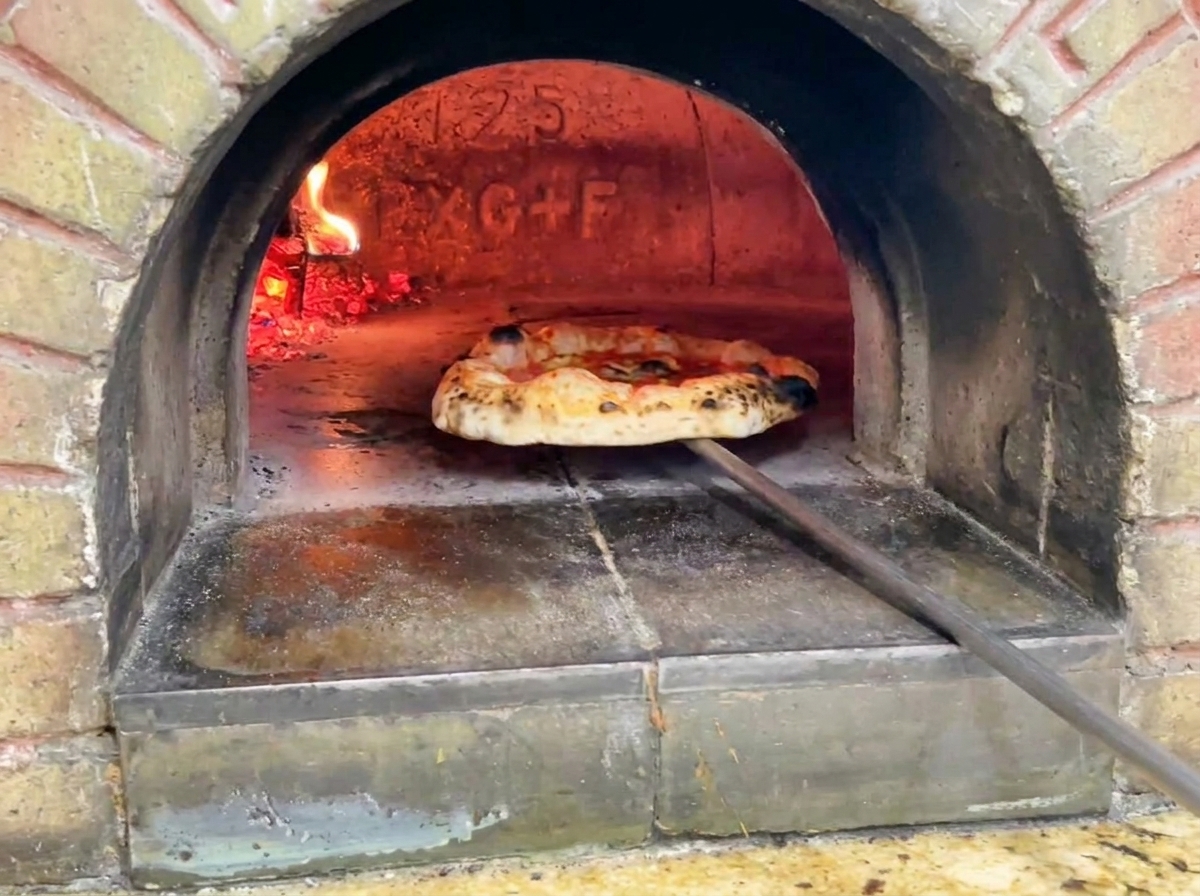 The finished pizza being pulled out of the brick oven showing a golden bubbly crust with dark charred spots