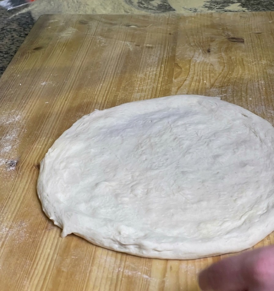 A chef spreading a layer of vibrant red tomato sauce over the center of a raw round pizza dough on a wooden board