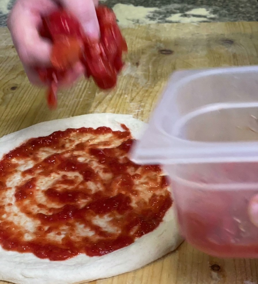 A hand dropping pieces of thick, crushed tomato pulp onto a pizza base already coated in smooth red sauce.