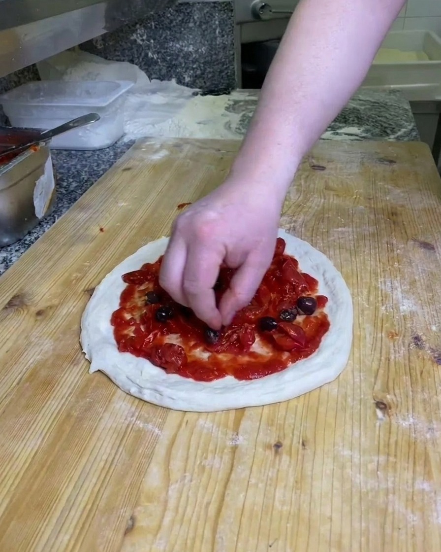 A hand placing dark, whole olives on top of the tomato-covered pizza dough on a floured wooden surface.