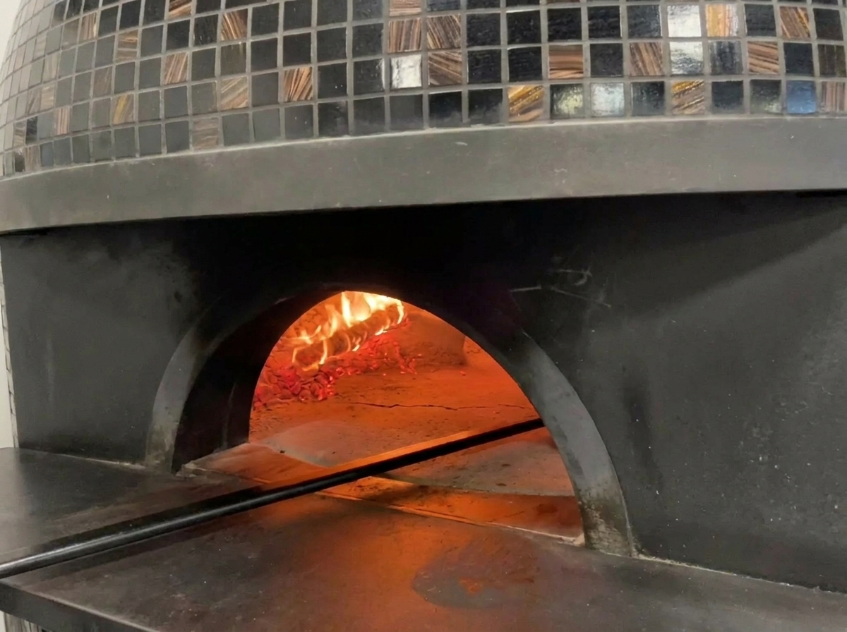 A view into the glowing mouth of a traditional tiled pizza oven with flames licking the dome and a pizza baking inside