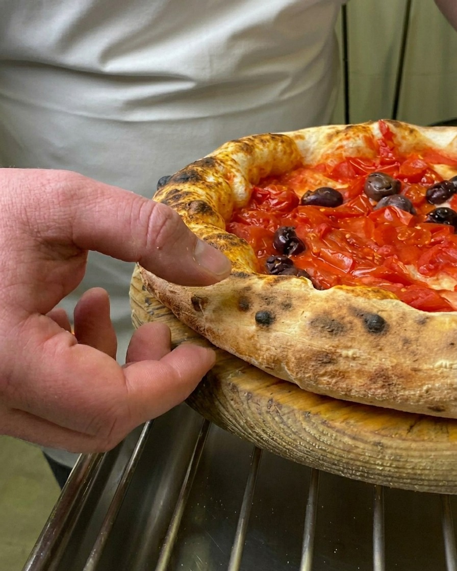 A chefs finger pressing into the soft, charred, and puffed-up crust of a freshly baked Marinara pizza.