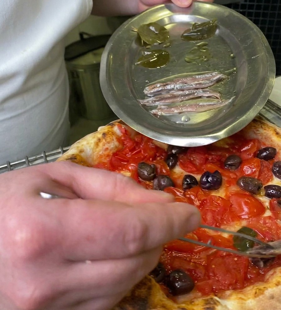 A chef using tweezers to pick up a pickled bay leaf from a metal plate to place it onto a pizza topped with tomatoes and olives.