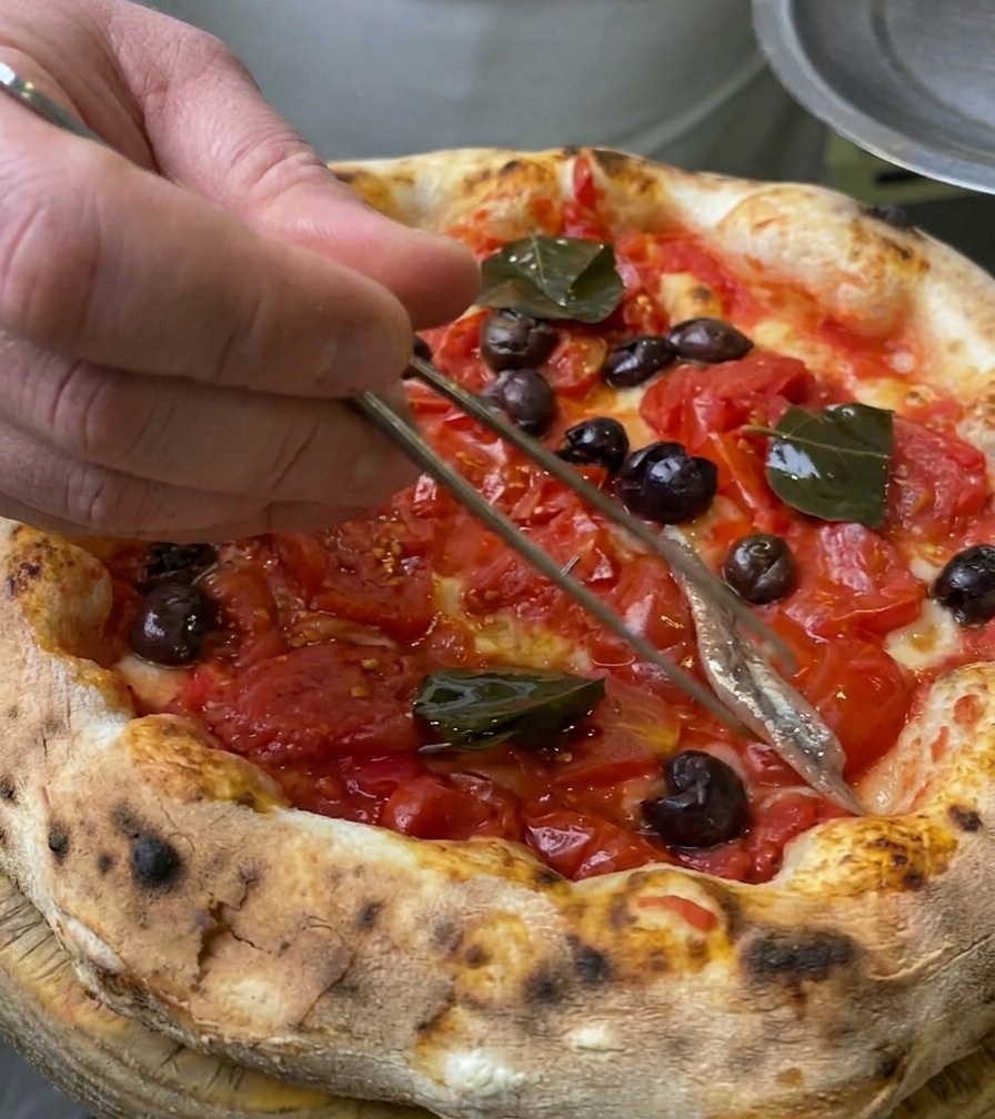 Close-up of a silver anchovy fillet being placed onto a pizza crust using metal culinary tweezers.