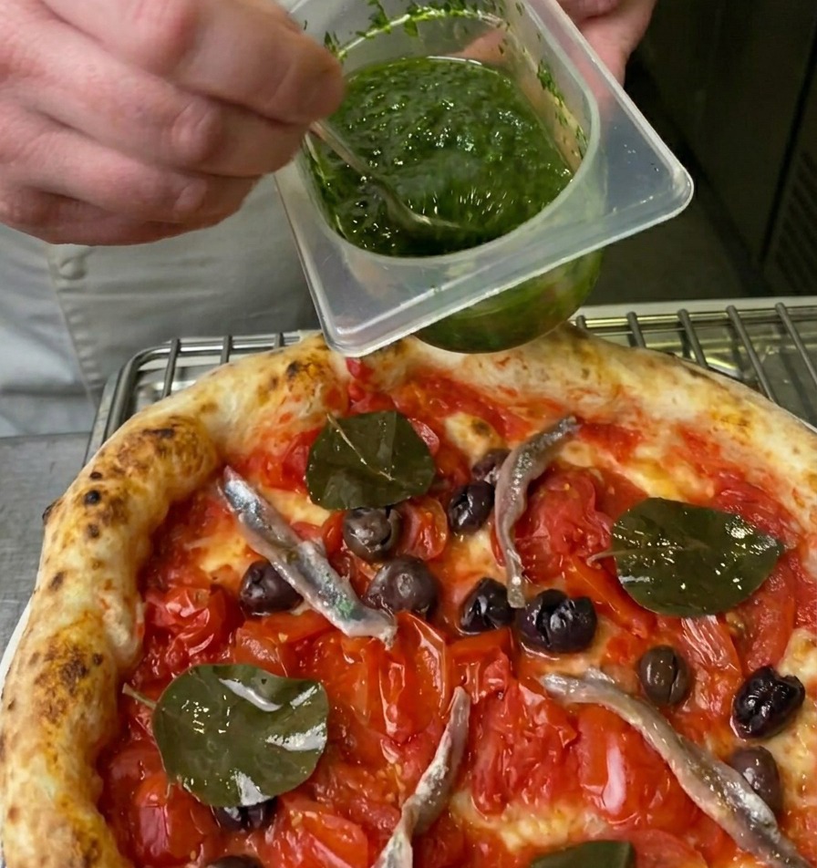 A chef drizzling a bright green herb sauce from a plastic container over a pizza using a small spoon.