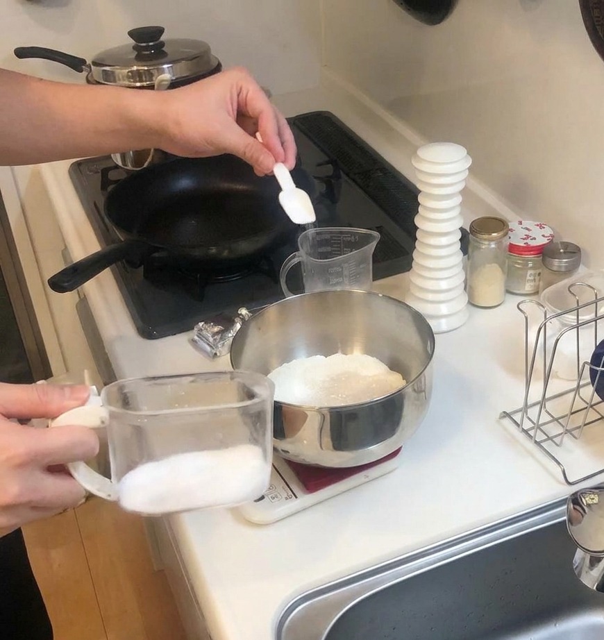 A small white scoop adding salt to a bowl of flour and yeast resting on a kitchen scale.