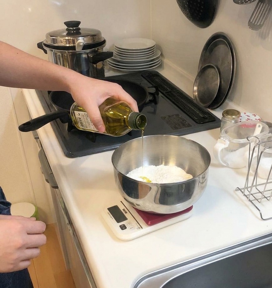 A stream of yellow olive oil being poured from a glass bottle into a metal mixing bowl containing dry flour.