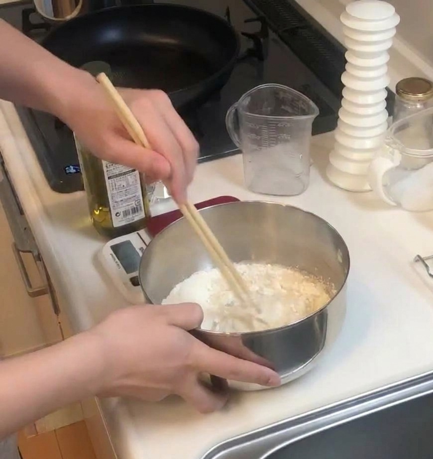 A person using two wooden chopsticks to vigorously stir a mixture of flour and liquids in a stainless steel bowl.