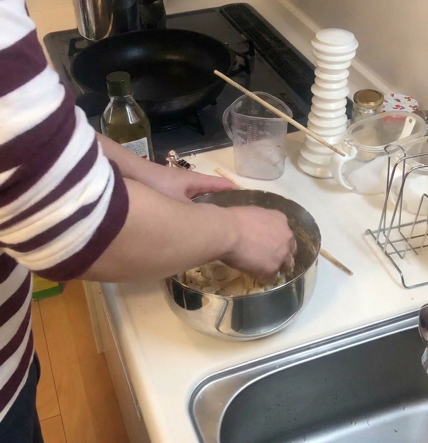 Hands kneading pizza dough inside a stainless steel bowl on a white kitchen counter next to a bottle of olive oil.