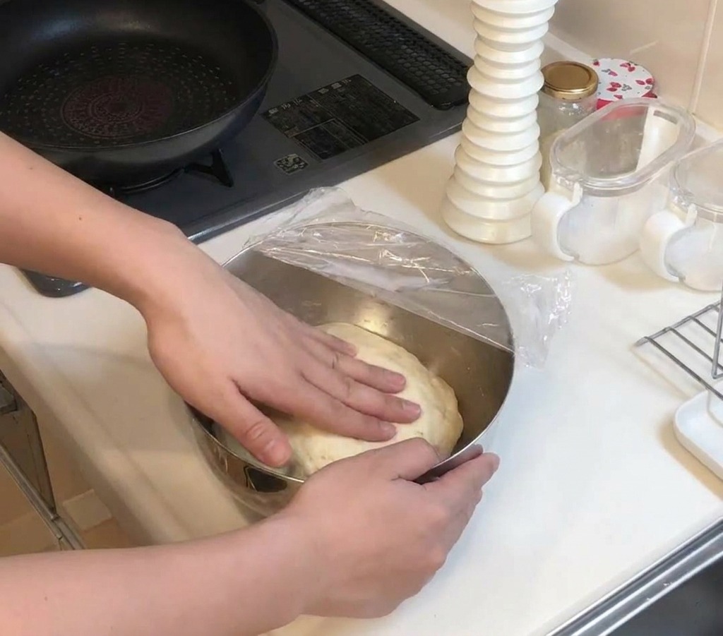 Hands pressing down on a large mound of risen, pale dough inside a metal bowl covered with tight plastic wrap.