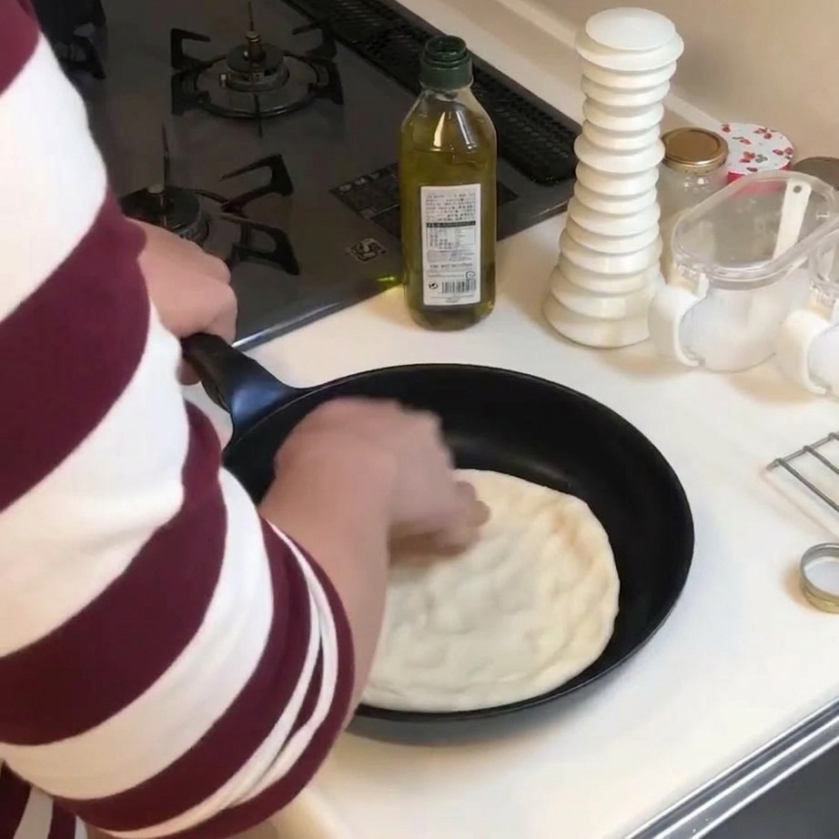 A person using their hands to press and flatten a circle of pizza dough into a black frying pan.
