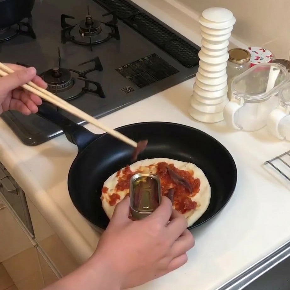 Using wooden chopsticks to pick up anchovies from a small metal tin and place them onto a sauced pizza base.